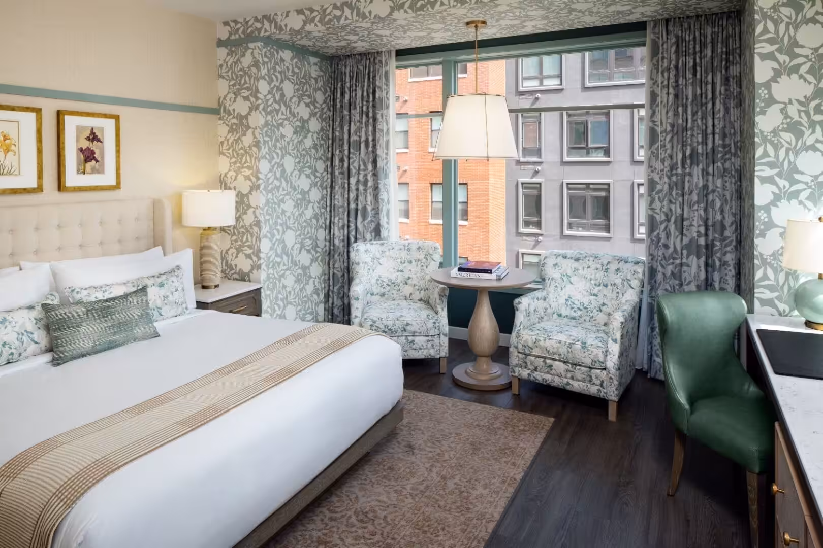 Elegant hotel room featuring a king bed, floral armchairs, and a large window with city views.